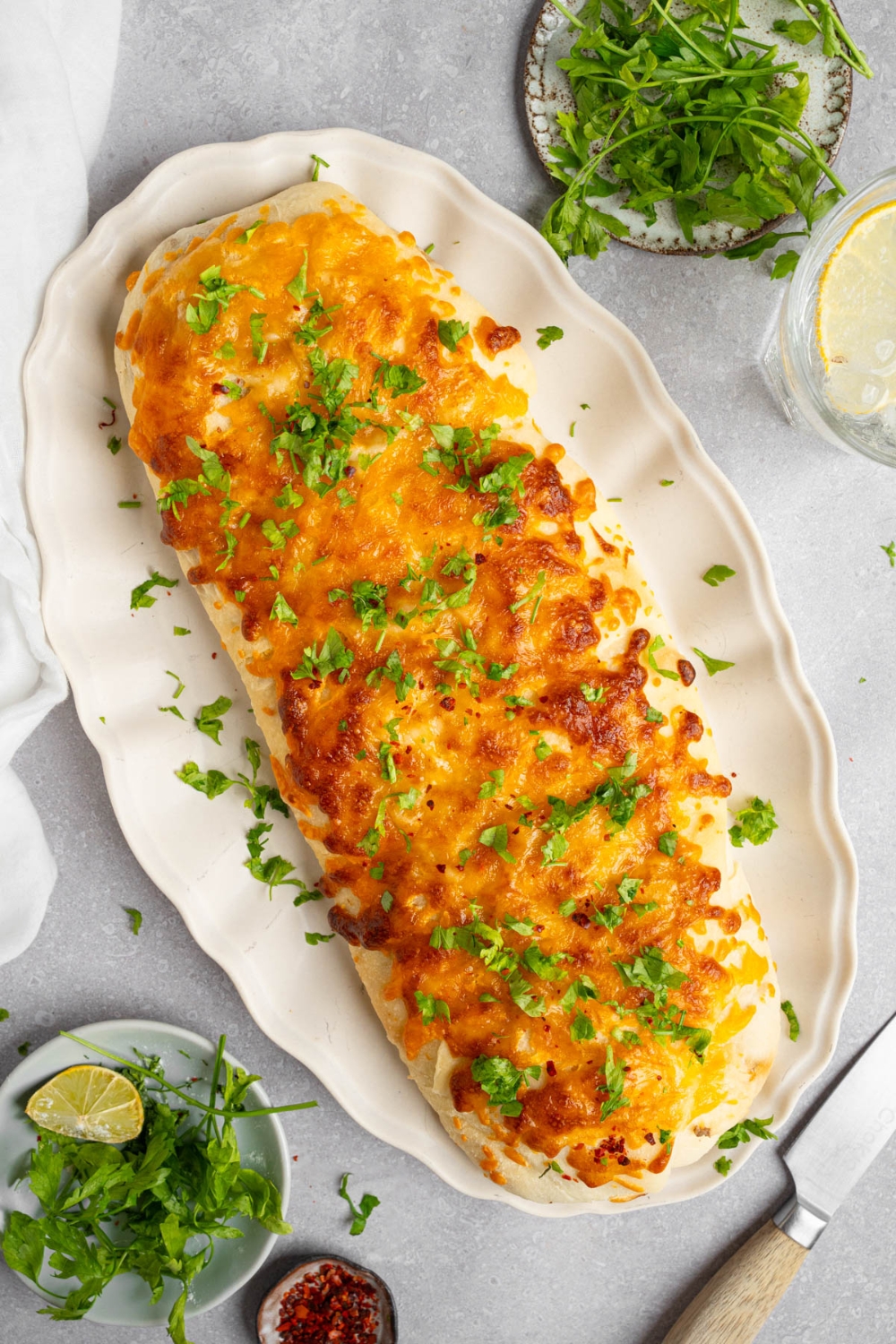 A loaf of garbage bread on a white platter garnished with chopped parsley. The platter is on a white counter with a glass of lemon water and small ramekins of garnishes.
