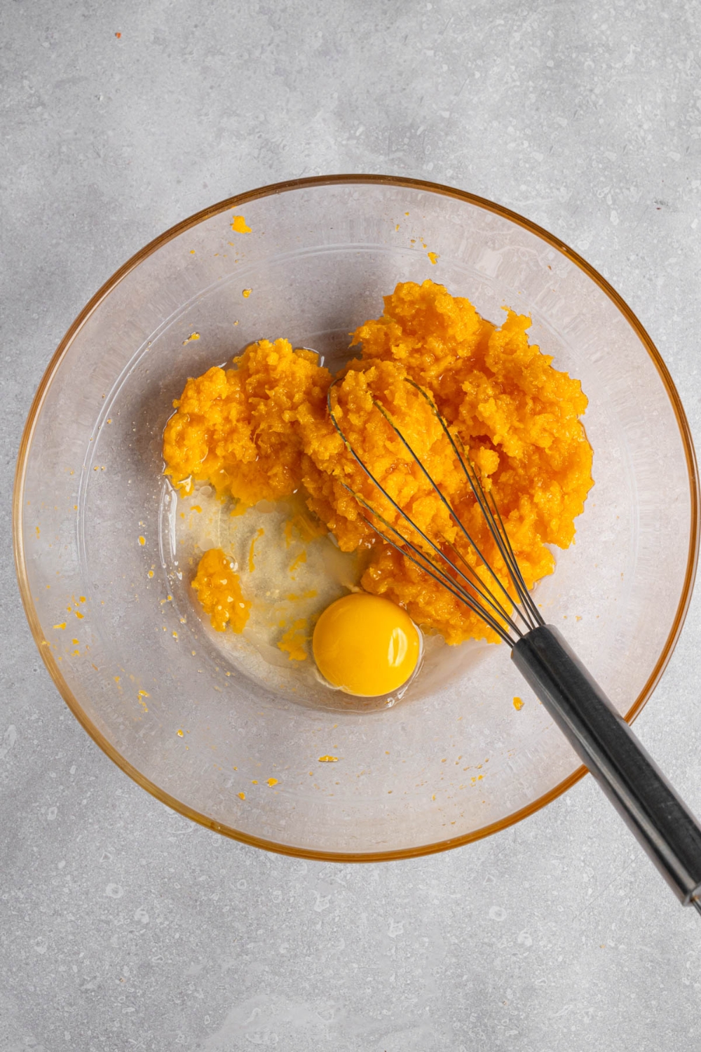 A glass bowl with a whisk mixing pumpkin puree, oil, and egg. The bowl is on a white counter.