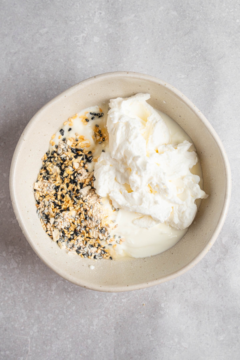 A white bowl with ingredients to make bagel dip including greek yogurt, cream cheese, and everything bagel seasoning. The bowl is on a white counter.