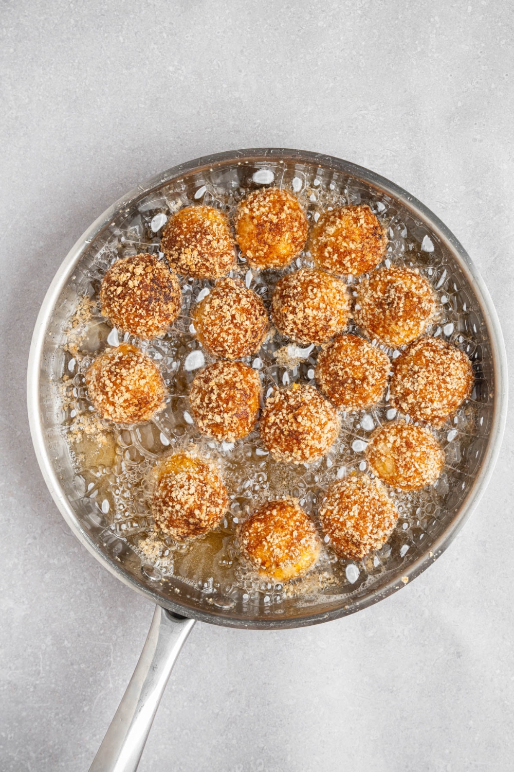 A skillet with breaded mashed potato balls frying in oil. The skillet is on a white counter.