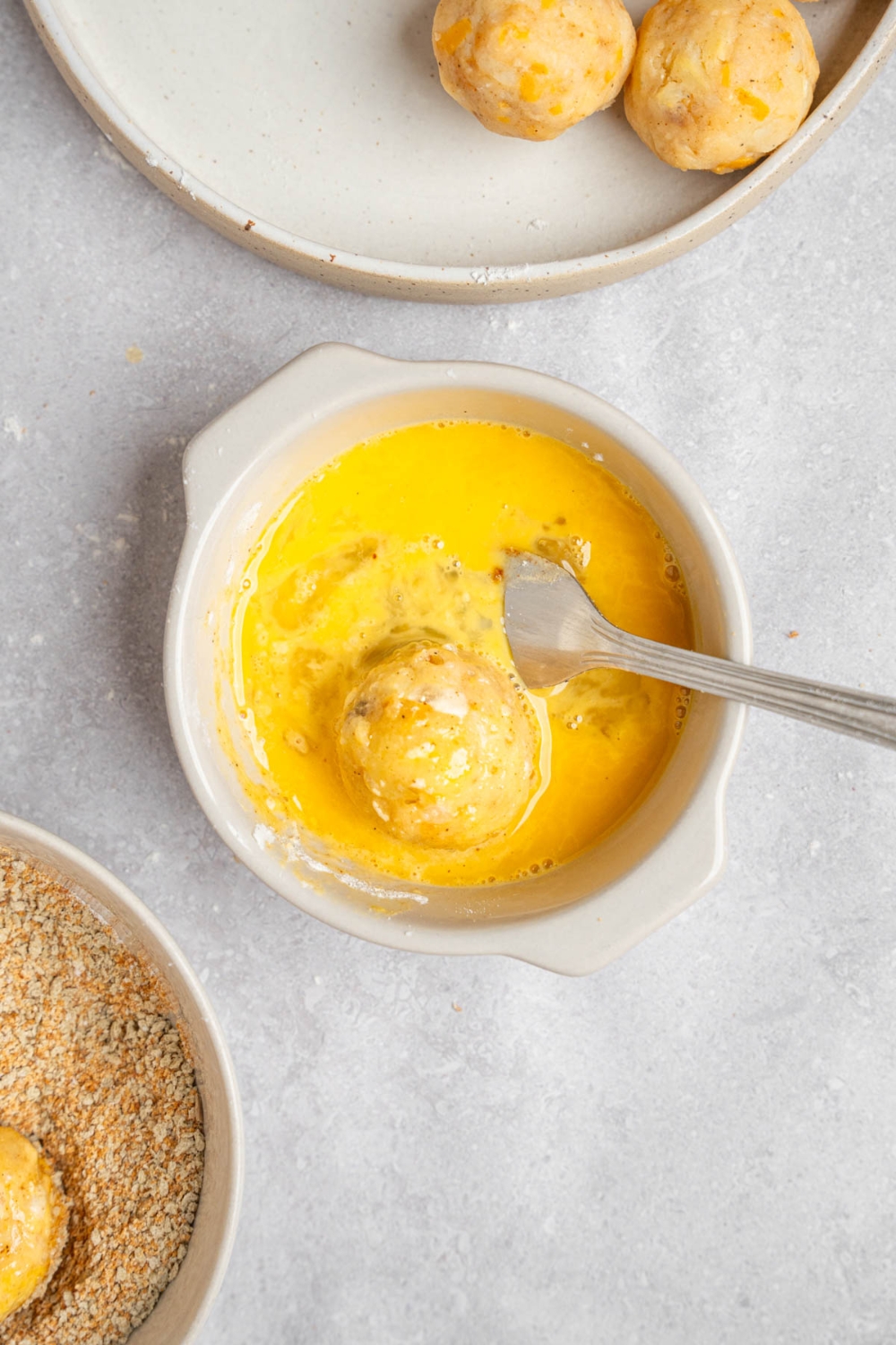 A small ramekin of whisked egg and a fork. There is an uncooked mashed potato ball in the egg mixture. The ramekin is on a white counter with a plate of uncooked mashed potato balls.
