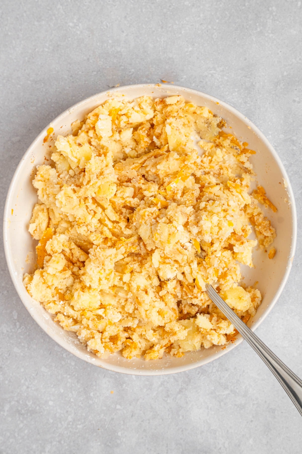 A white bowl with a fork mixing a cheesy mashed potato mixture. The bowl is on a white counter.