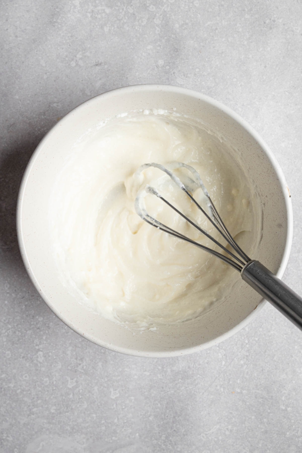 A white bowl with a whisk mixing cream cheese filling. The bowl is on a white counter.