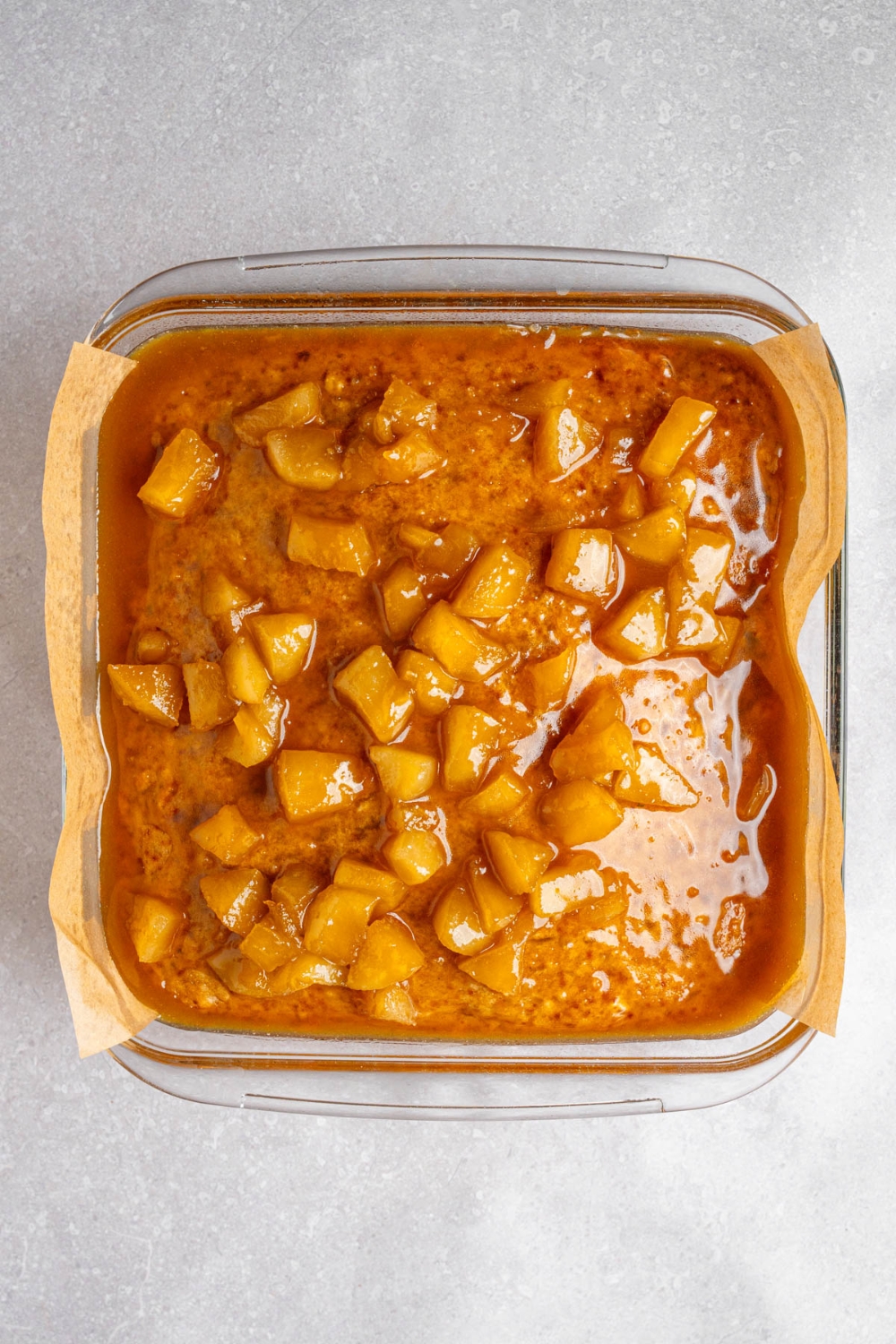 A glass baking dish lined with parchment paper with cooked Amish applesauce cake topped with a caramel apple glaze. The dish is on a white counter.