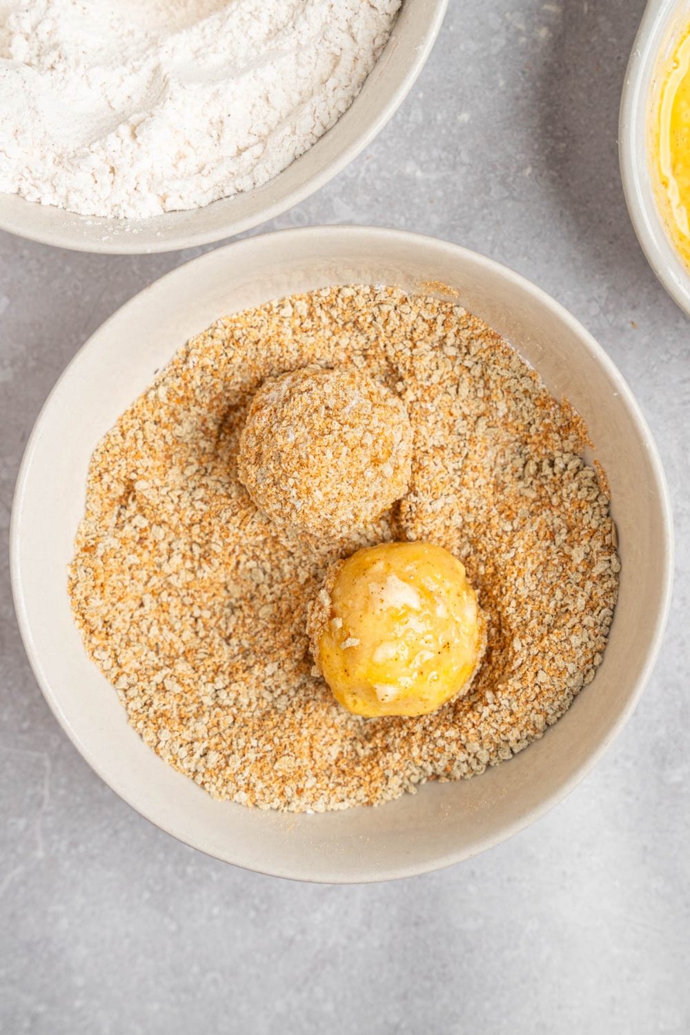 A white bowl with two uncooked mashed potato balls in a breadcrumb mixture. The bowl is on a white counter with a bowl of flour and bowl of egg.