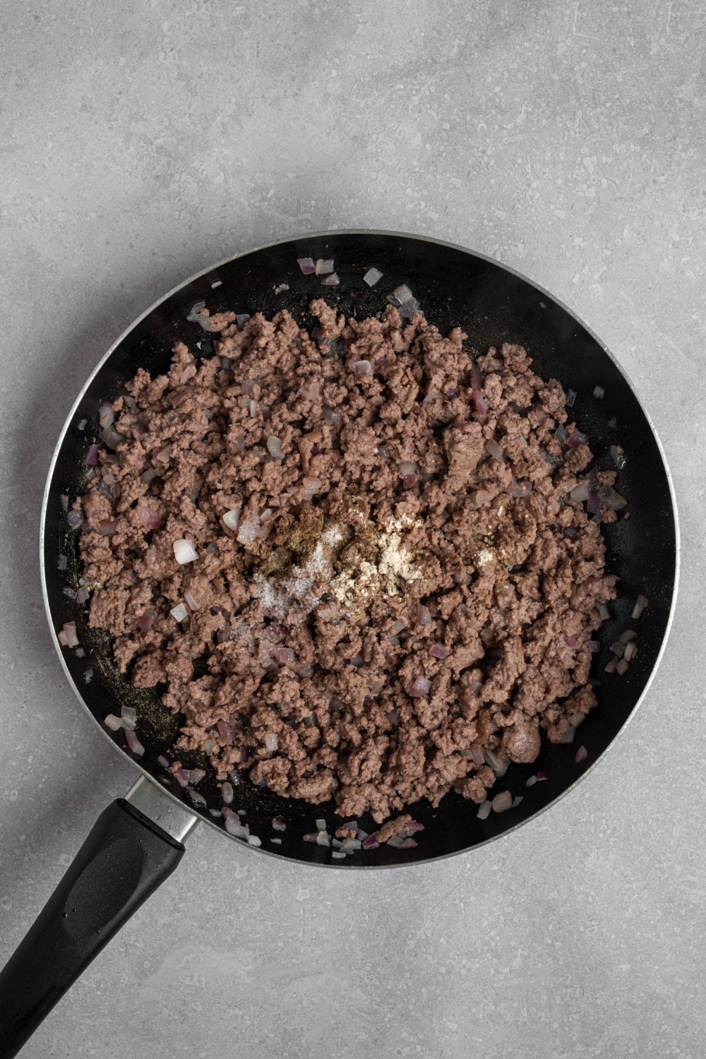 A skillet with cooked ground beef topped with seasonings. The skillet is n a white counter.