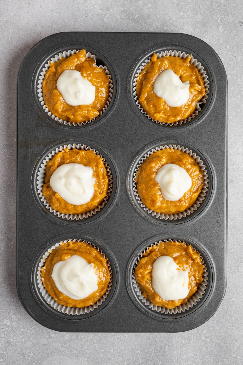 A muffin tray with six uncooked copycat Starbucks pumpkin muffins topped with cream cheese filling. The tray is on a white counter.