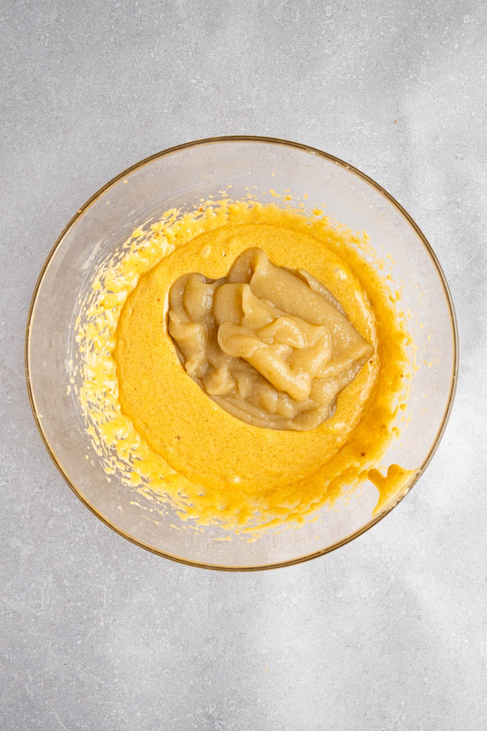 A glass bowl with wet ingredients combined to make Amish applesauce cake with applesauce on top. The bowl is on a white counter