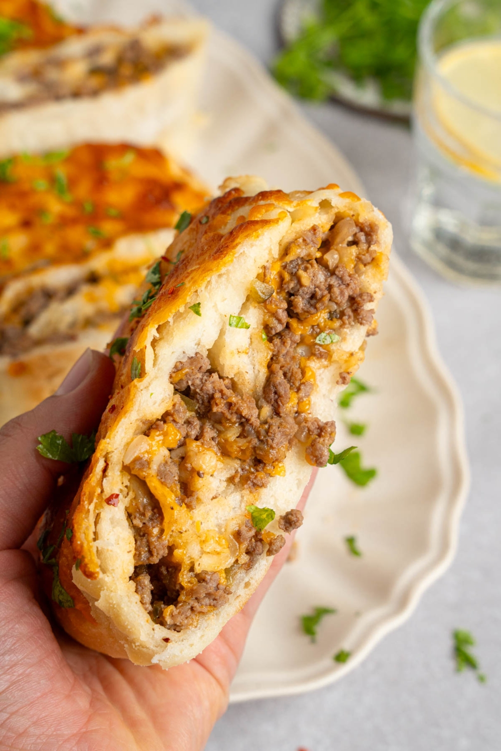 A hand holding a slice of garbage bread garnished with fresh parsley. There is a platter of sliced garbage bread blurred in the background.