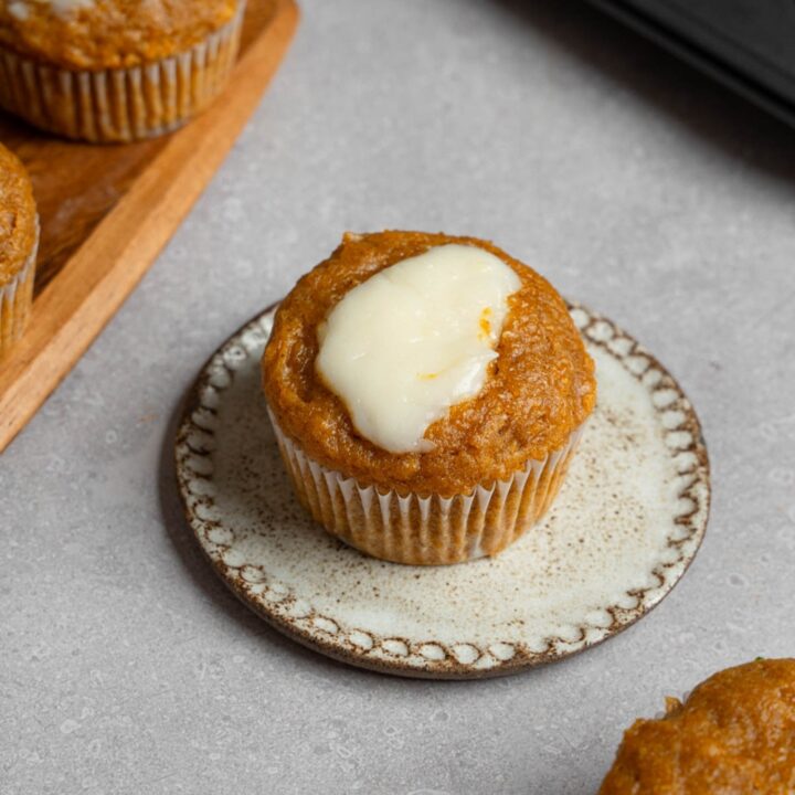 A homemade Starbucks pumpkin muffin topped with cream cheese filling on a small ceramic plate. The plate is on a white counter with a tray of baked pumpkin muffins.