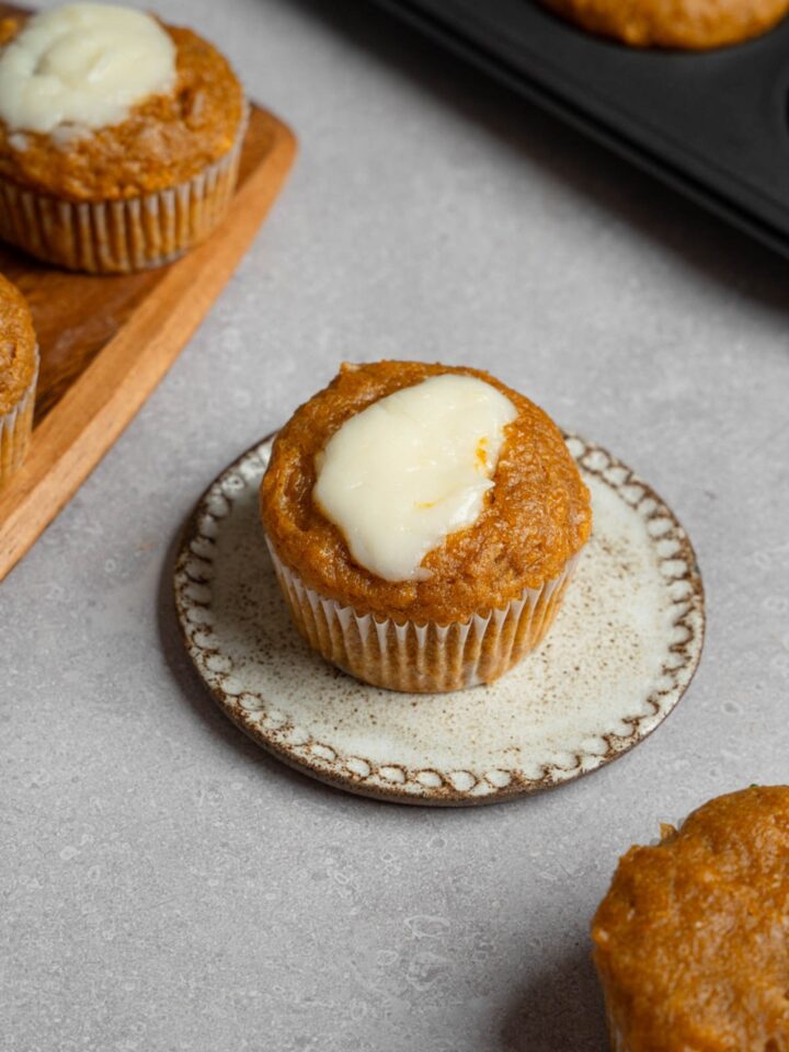 A homemade Starbucks pumpkin muffin topped with cream cheese filling on a small ceramic plate. The plate is on a white counter with a tray of baked pumpkin muffins.