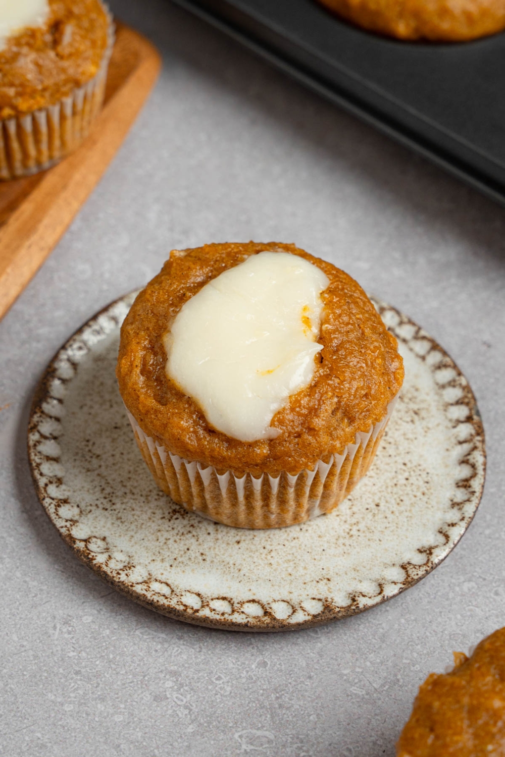 A homemade Starbucks pumpkin muffin topped with cream cheese filling on a small ceramic plate. The plate is on a white counter with a tray of baked pumpkin muffins.