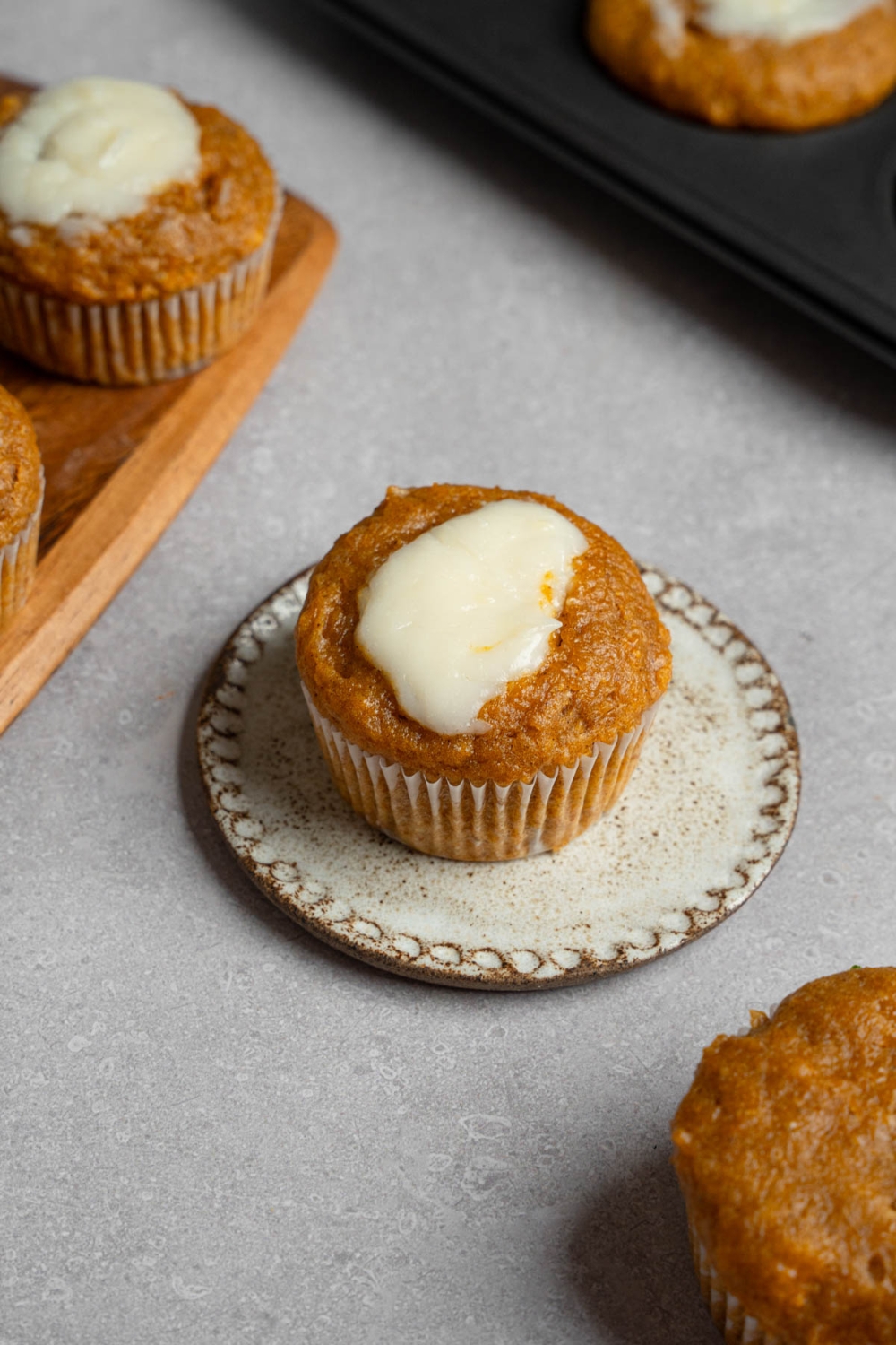 A homemade Starbucks pumpkin muffin topped with cream cheese filling on a small ceramic plate. The plate is on a white counter with a tray of baked pumpkin muffins.