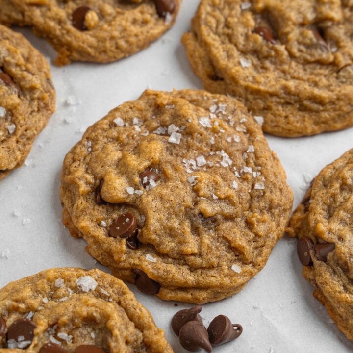 Banana bread cookies sprinkled with flaky sea salt on a baking sheet lined with parchment paper. The baking sheet is garnished with chocolate chips.