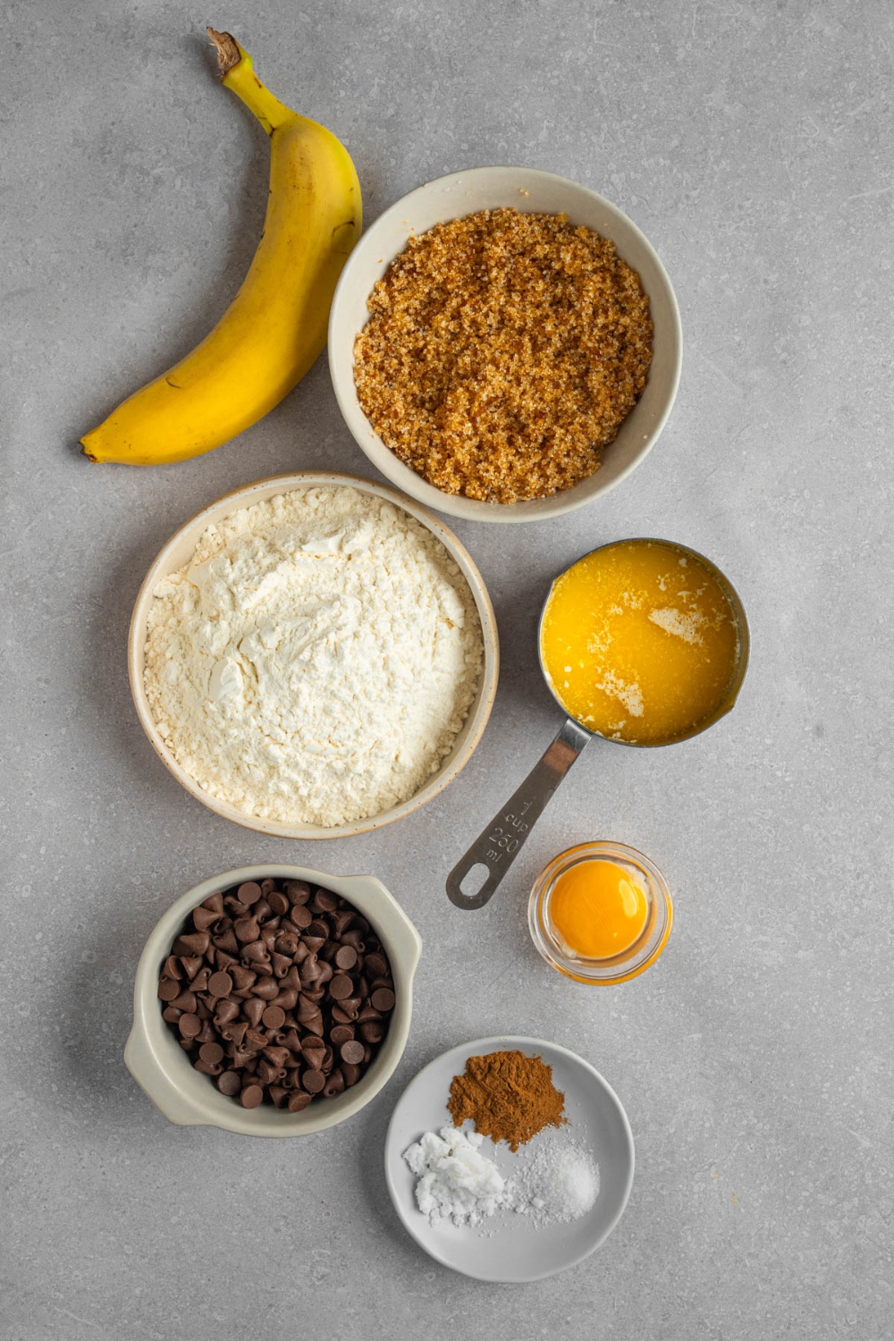 An overhead shot of several bowls of various sizes containing ingredients to make banana bread cookies including flour, banana, cinnamon, brown sugar, chocolate chips, butter, and egg.