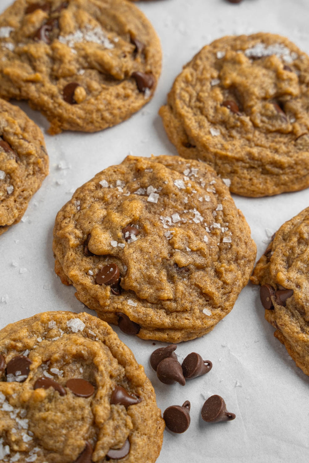 Banana bread cookies sprinkled with flaky sea salt on a baking sheet lined with parchment paper. The baking sheet is garnished with chocolate chips.