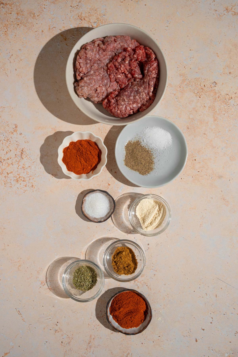 An overhead shot of several bowls in various sizes containing ingredients to make beef chorizo including ground beef, salt, paprika, ancho chile powder, cumin, oregano, garlic powder, and other spices.