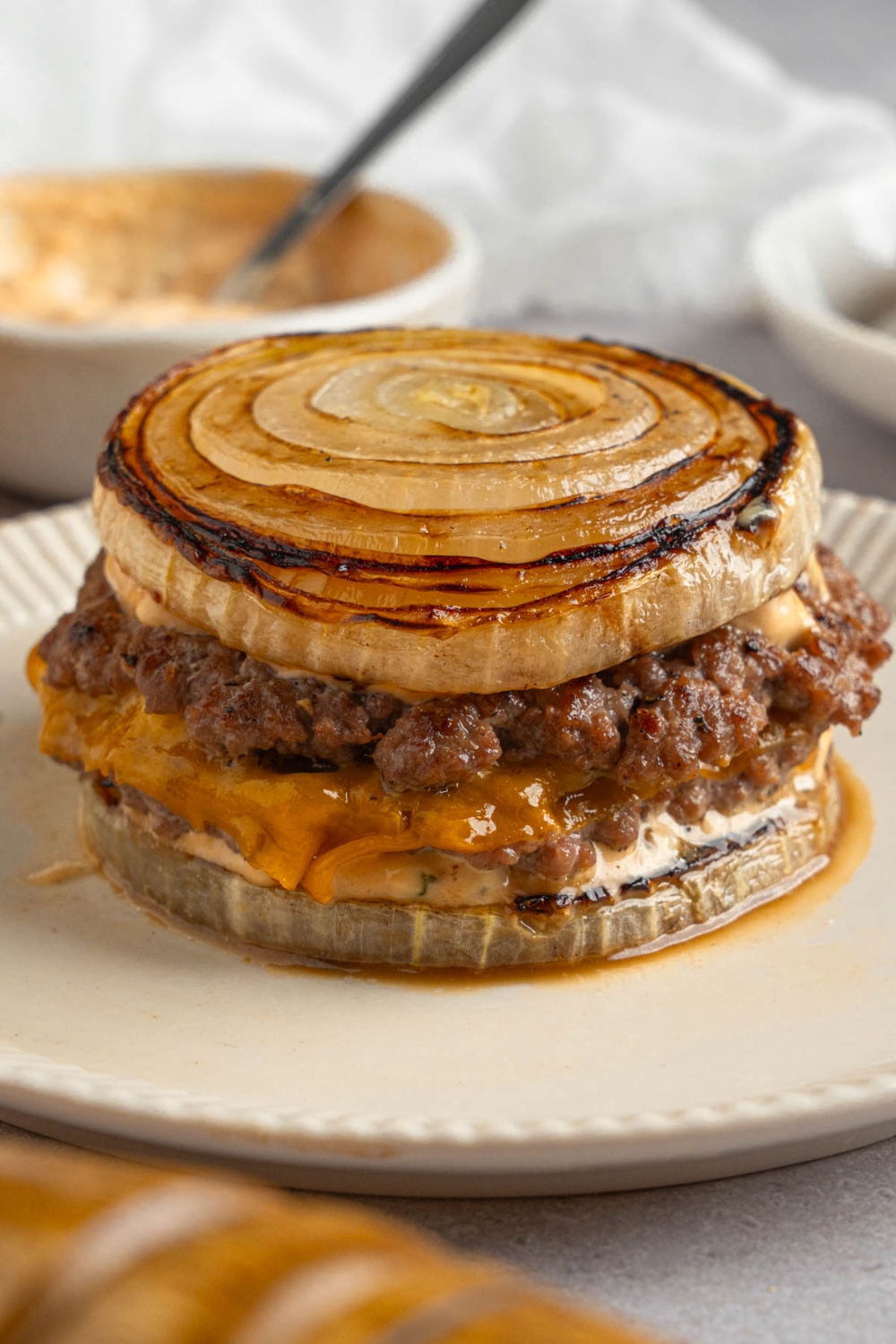 A Flying Dutchman burger in between grilled onions on a white plate. The plate is on a white counter with a bowl of burger sauce with a spoon.