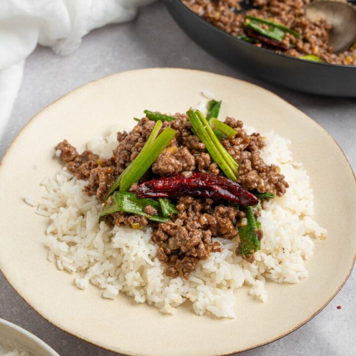 A white plate with Mongolian ground beef with green onion and dried chilis served over white rice. The plate is on a white counter with a skillet of ground beef and bowl of white rice.