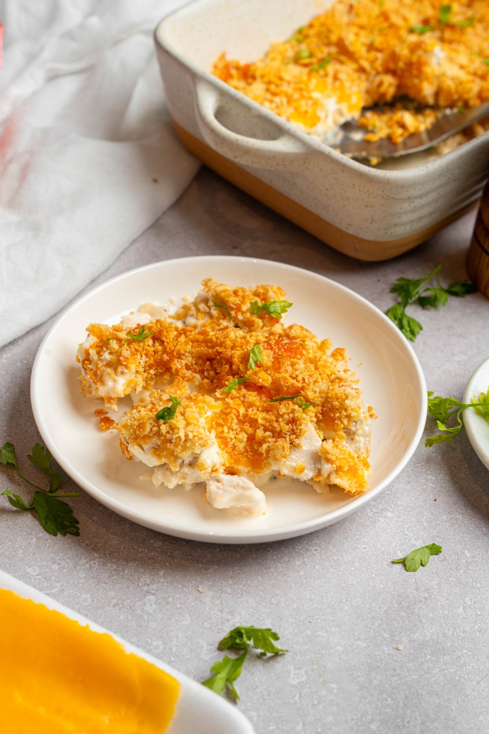 A plate of Neiman Marcus chicken casserole garnished with fresh parsley. The plate is on a white counter with a baking dish of casserole and a white cloth napkin.