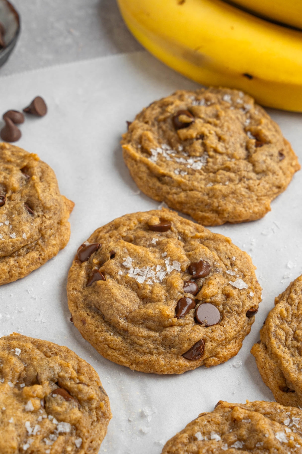 Banana bread cookies sprinkled with flaky sea salt on parchment paper. The parchment paper is garnished with chocolate chips and banana.