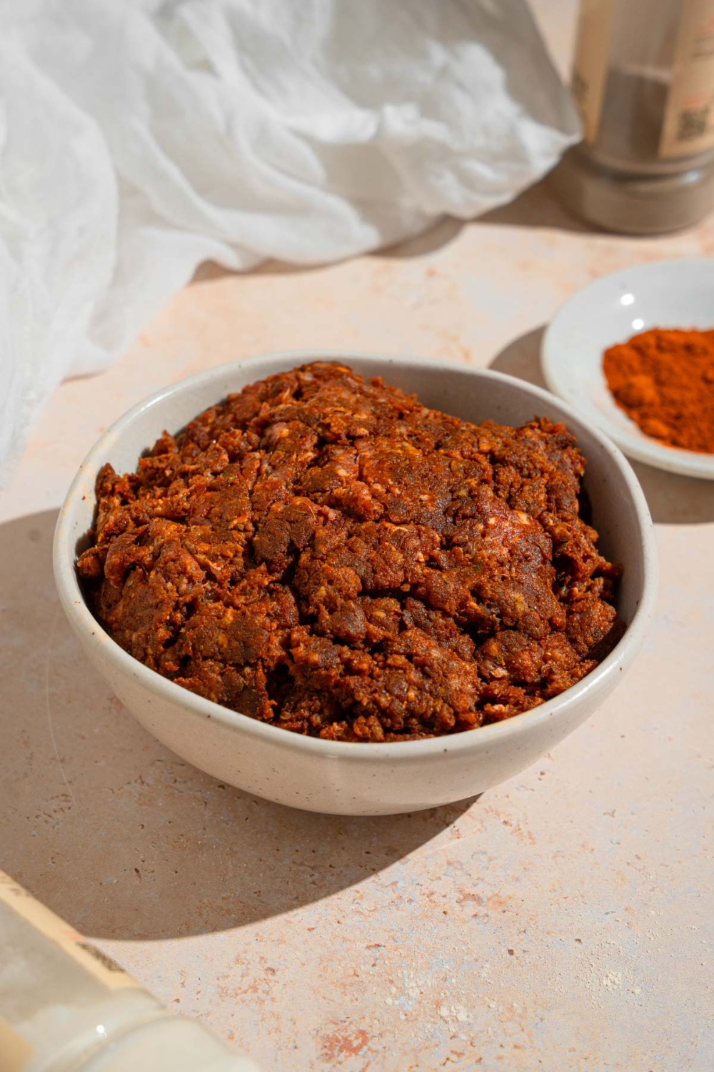 Beef chorizo in a white bowl on a tan counter with a white cloth napkin and small plate of spices.