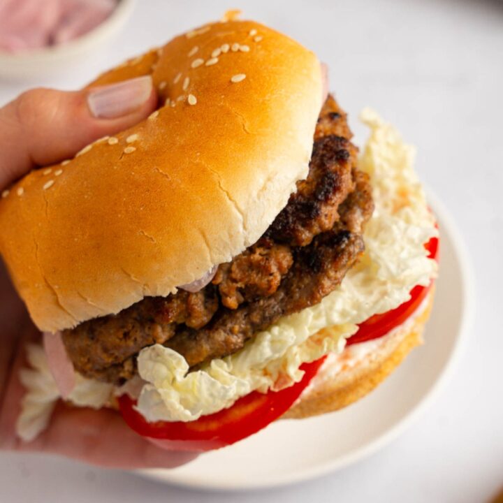 A close up of a hand holding a brisket burger with sliced tomato. There is a white plate on a white counter blurred in the background.