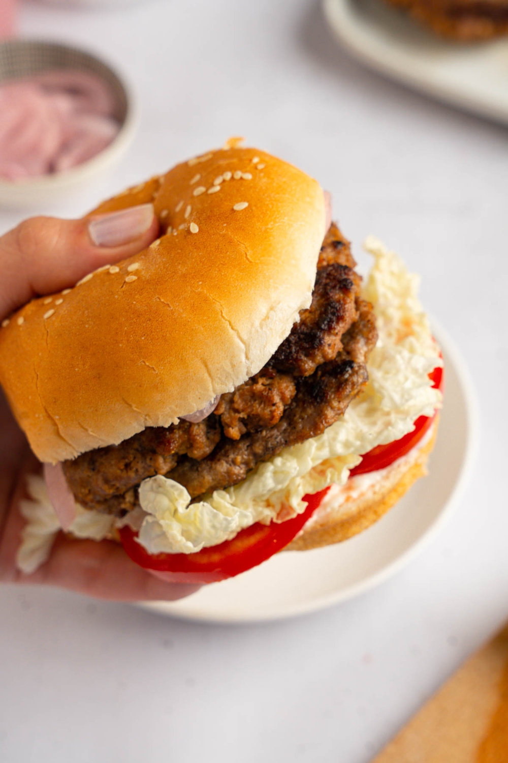 A close up of a hand holding a brisket burger with sliced tomato. There is a white plate on a white counter blurred in the background.