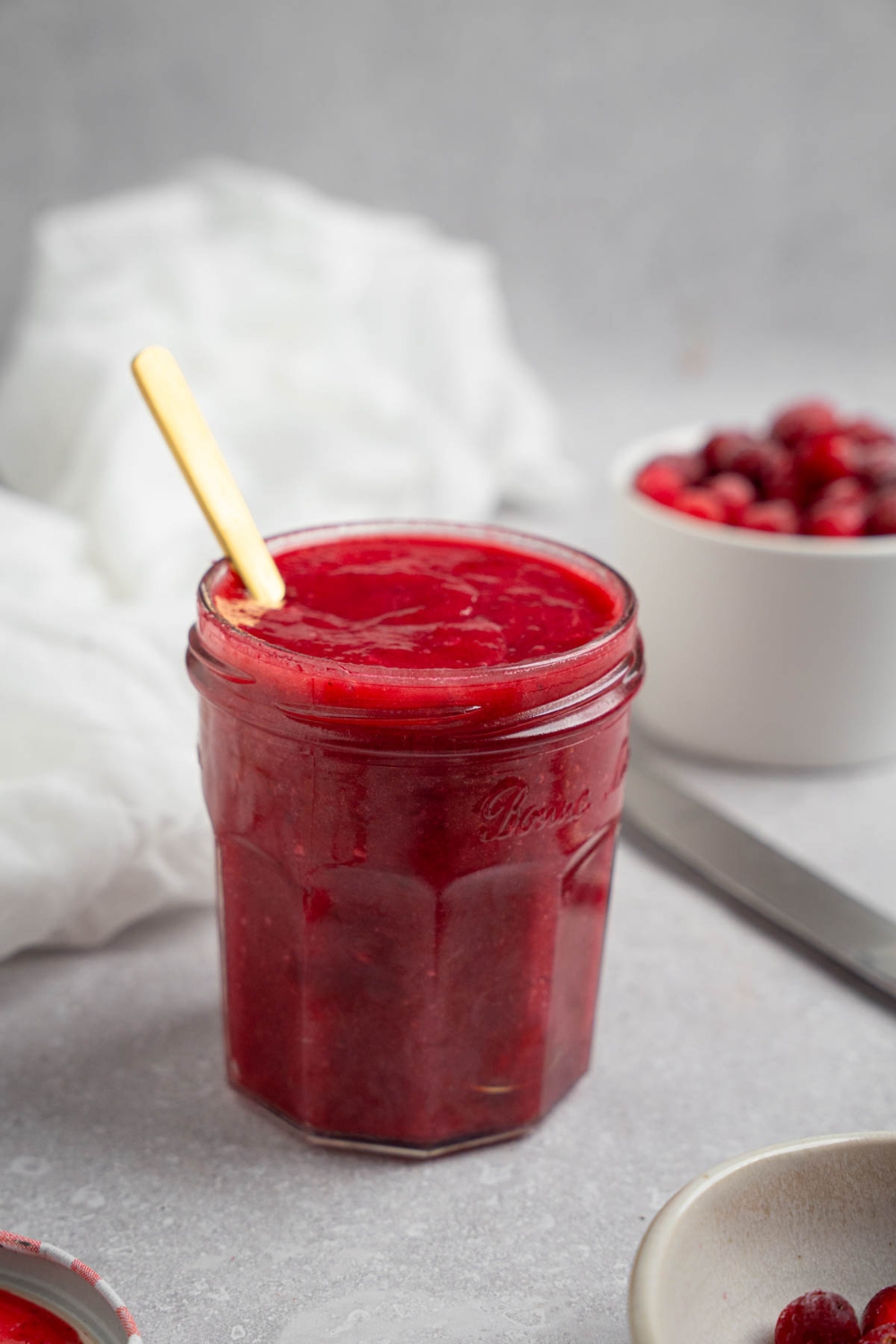 A glass mason jar of chokecherry pudding with a spoon. The jar is on a white counter with a white cloth napkin and white bowl of chokecherries.