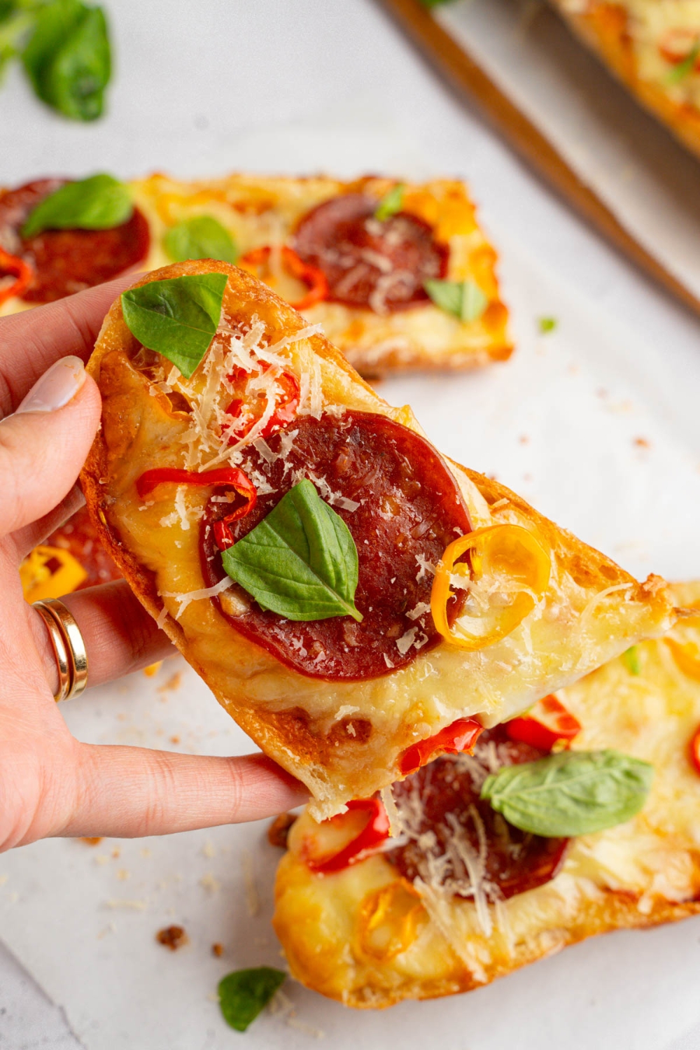 A close up of a hand holding a piece of garlic bread pizza garnished with fresh basil, shaved parmesan, and sliced sweet pepper. There are additional pieces of garlic bread pizza on a piece of parchment paper blurred in the background.