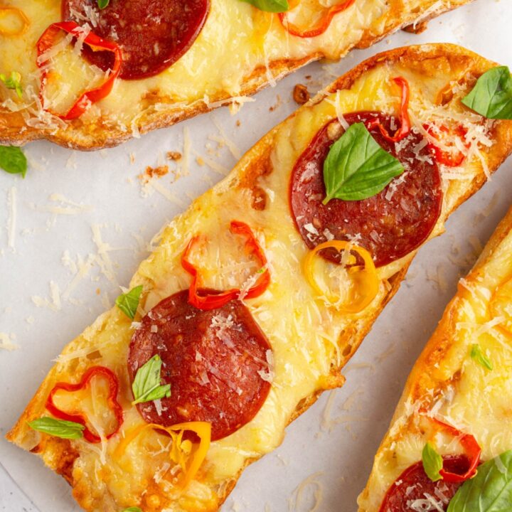 Three pieces of garlic bread pizza topped with pepperoni, sliced sweet peppers, gresh basil, and shaved parmesan. The pizza is on a piece of parchment paper.