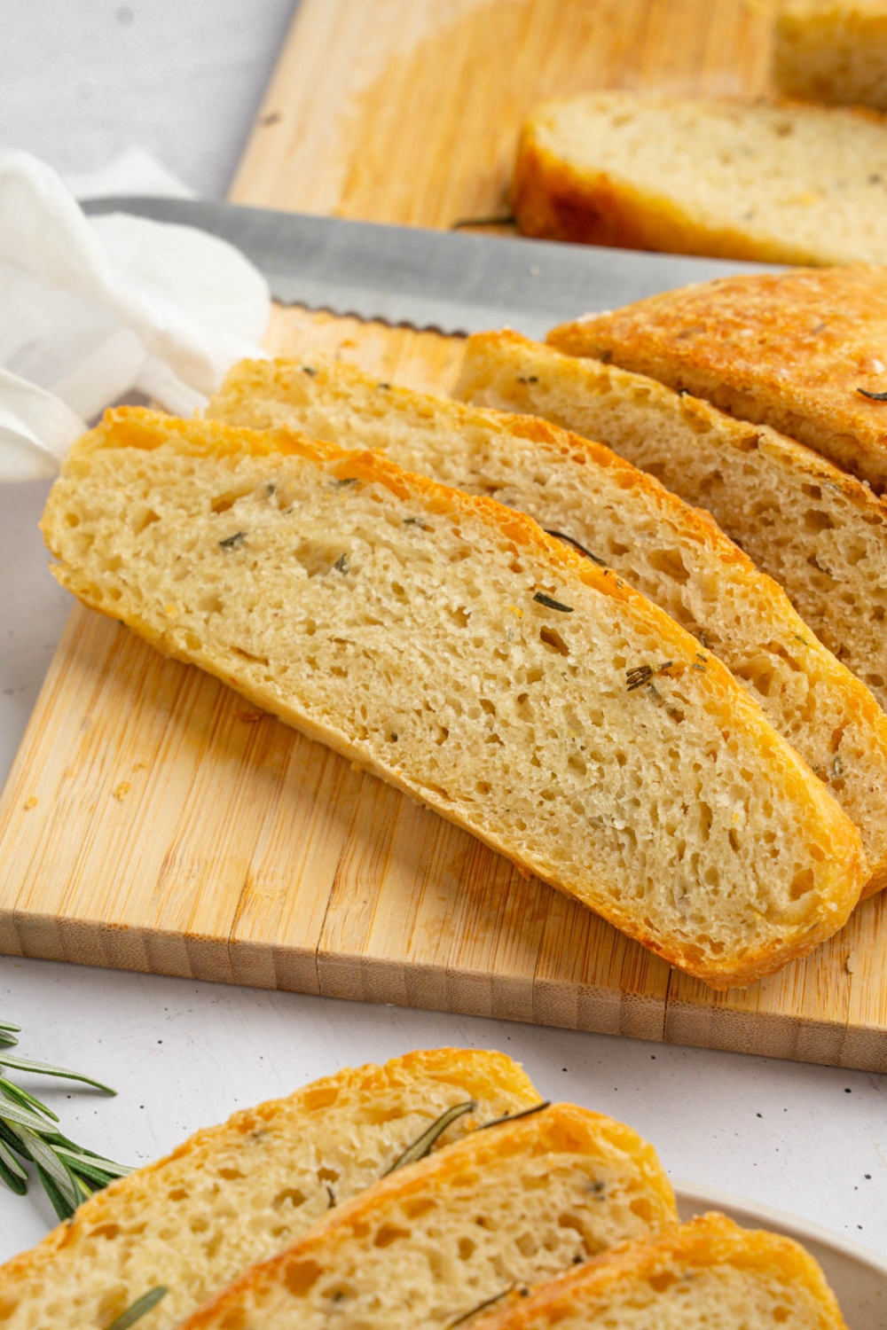 Several slices of garlic rosemary bread on a wooden board garnished with fresh rosemary. The board is on a white counter with a white cloth napkin.