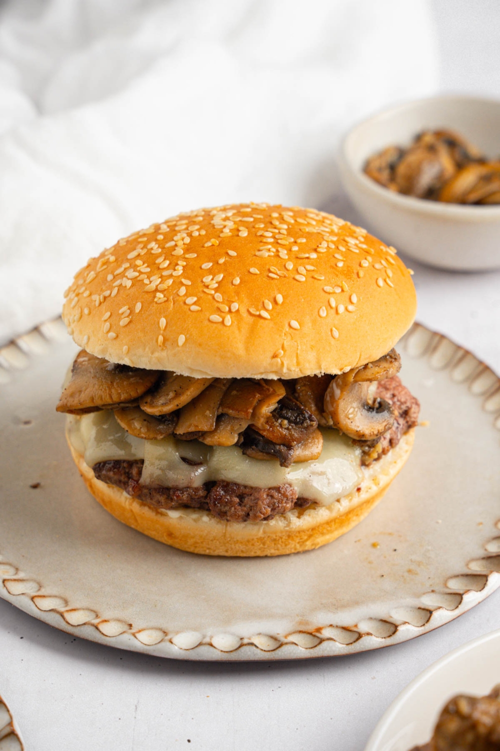 A mushroom swiss burger on a ceramic plate. The plate is on a white counter with a bowl of sauteed mushrooms and white cloth napkin.