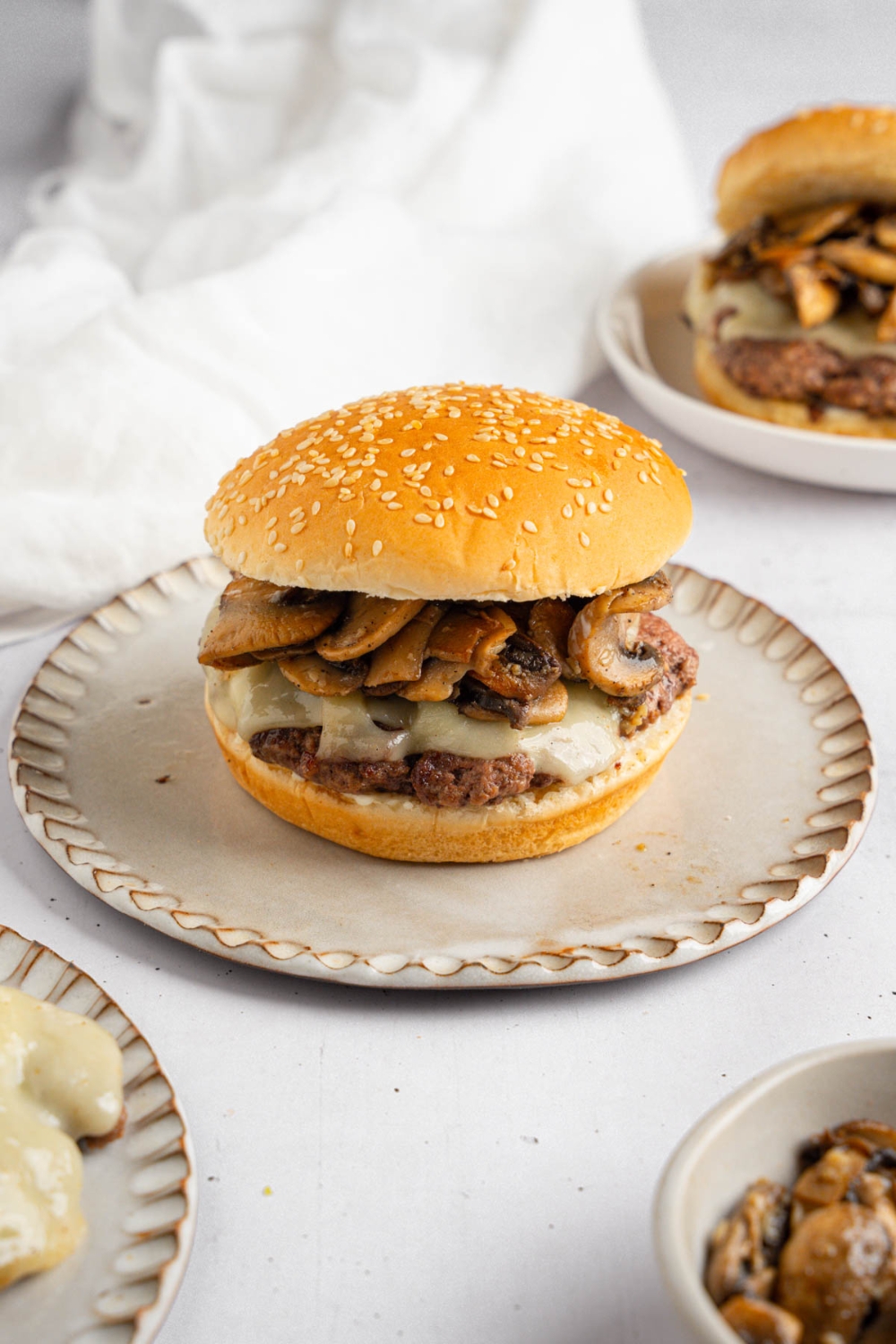 A mushroom swiss burger on a ceramic plate. The plate is on a white counter with a bowl of sauteed mushrooms, white cloth napkin, and an additional burger.