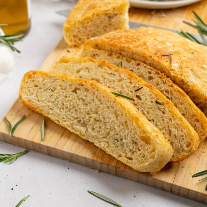Several slices of garlic rosemary bread on a wooden board garnished with fresh rosemary. The board is on a white counter with a white cloth napkin and jar of olive oil.