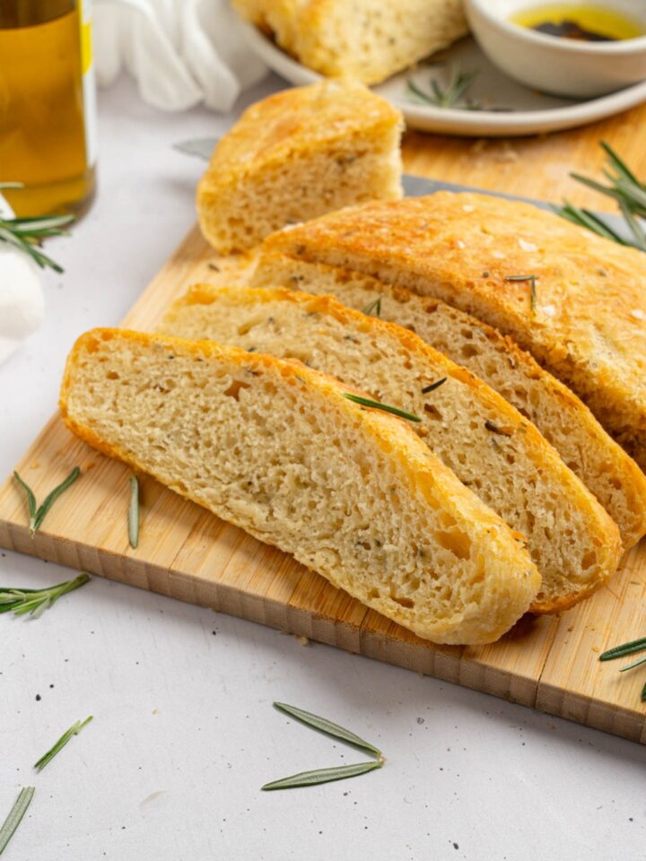 Several slices of garlic rosemary bread on a wooden board garnished with fresh rosemary. The board is on a white counter with a white cloth napkin and jar of olive oil.