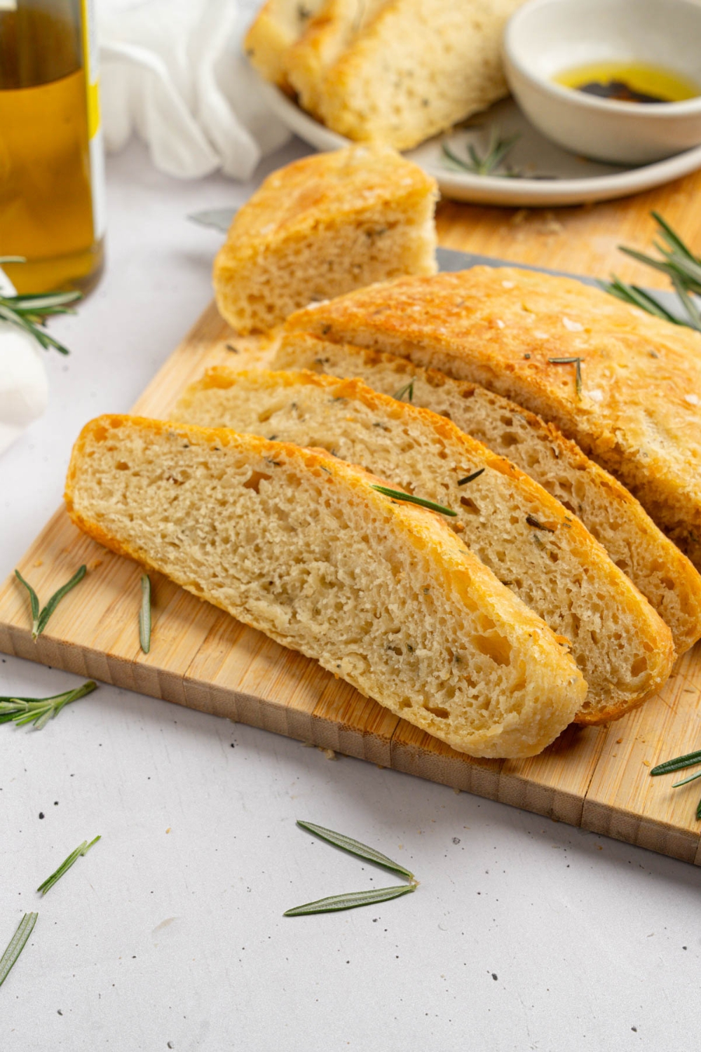 Several slices of garlic rosemary bread on a wooden board garnished with fresh rosemary. The board is on a white counter with a white cloth napkin and jar of olive oil.
