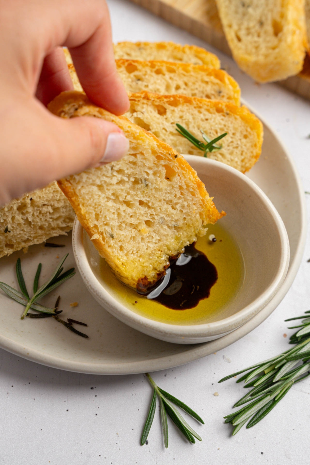A hand dipping a piece of garlic rosemary bread into a small bowl of oil and balsamic vinegar. The bowl is on a a plate with additional slices of bread garnished with fresh rosemary.