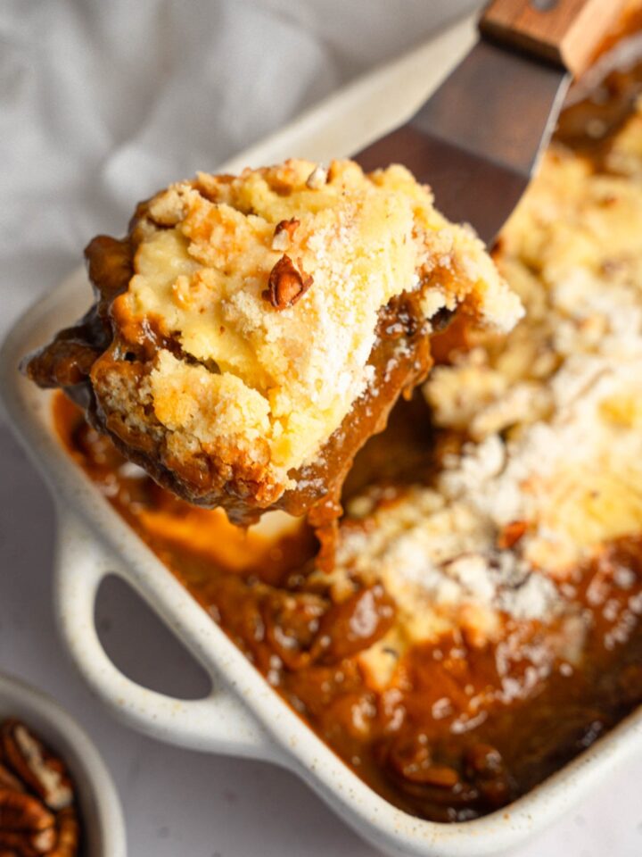 A close up of a spatula with a piece of pecan pie dump cake. There is a baking dish of dump cake blurred in the background.