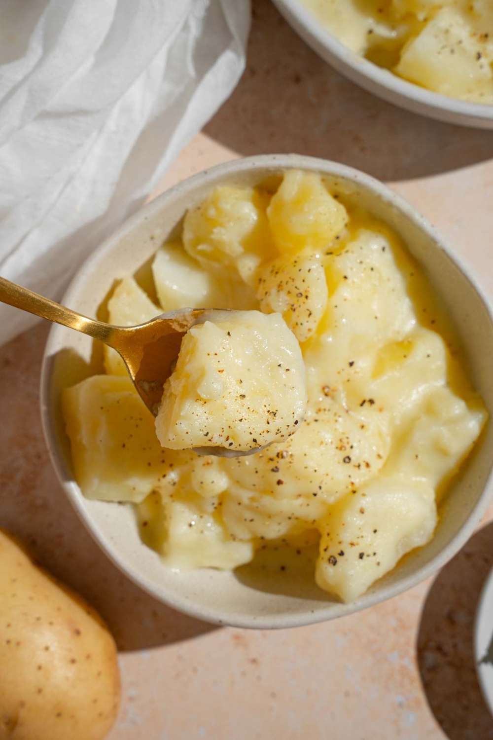 A bowl of stewed potatoes finished with ground pepper. A spoon is taking a bite of potatoes from the bowl. The bowl is on a tan counter.
