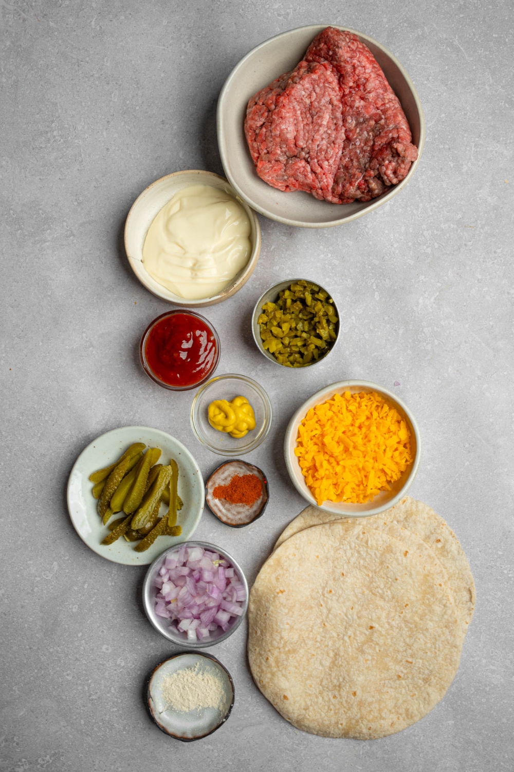 An overhead shot of several bowls in various sizes containing ingredients to make Big Mac wraps including ground beef, wraps, shredded cheese, sliced pickles, chopped red onion, ketchup, mustard, relish, mayo, and seasonings.