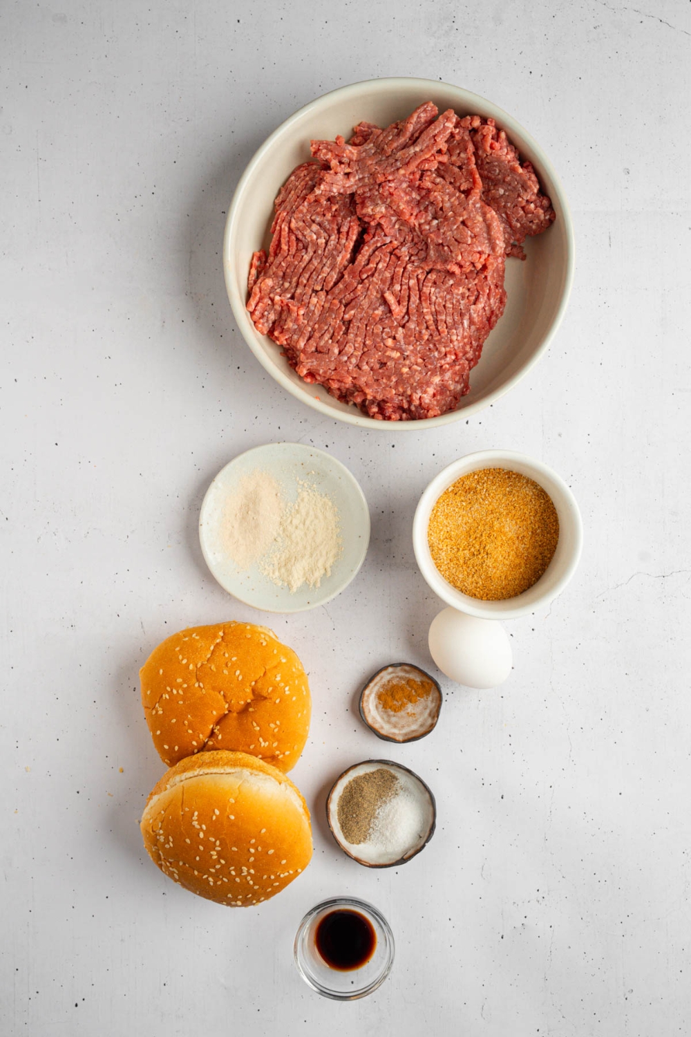 An overhead shot of several bowls in various sizes containing ingredients to make brisket burgers including ground brisket, breadcrumbs, worcestershire sauce, egg, hamburger buns, and seasonings.