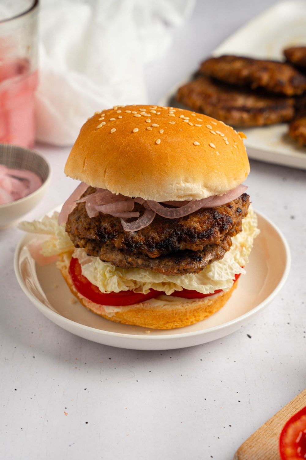 A brisket burger with sliced tomato, lettuce, and onion on a white plate. The plate is on a white counter with a tray of brisket patties and small ramekin of sliced onion.