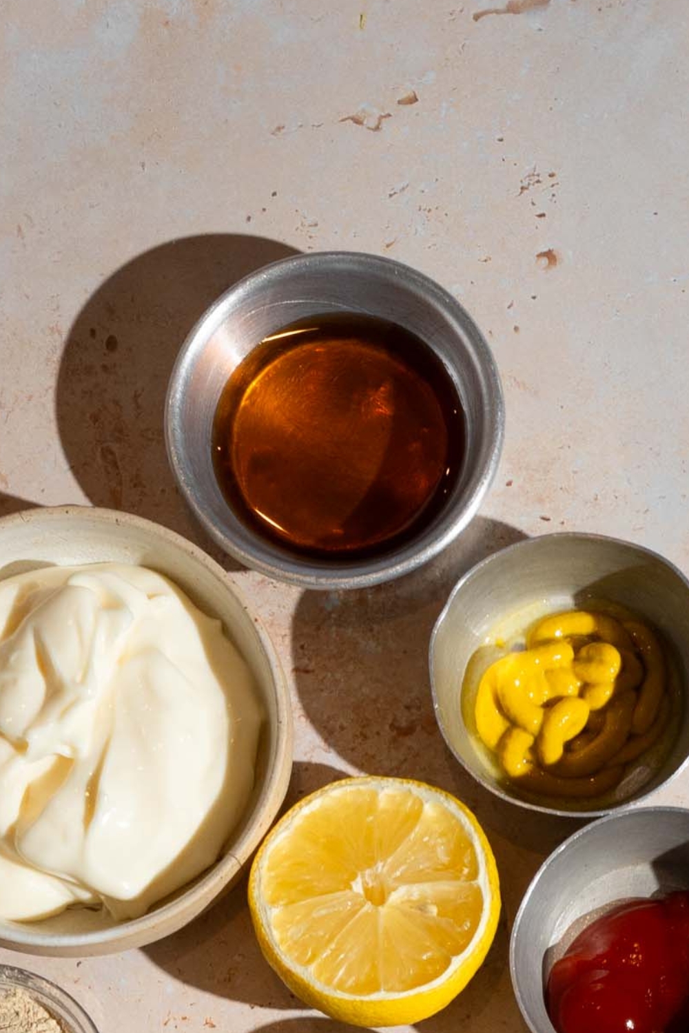 A bowl of honey, a bowl of mayonnaise, a bowl of mustard, half of lemon, and a bowl of barbecue sauce on a white counter.