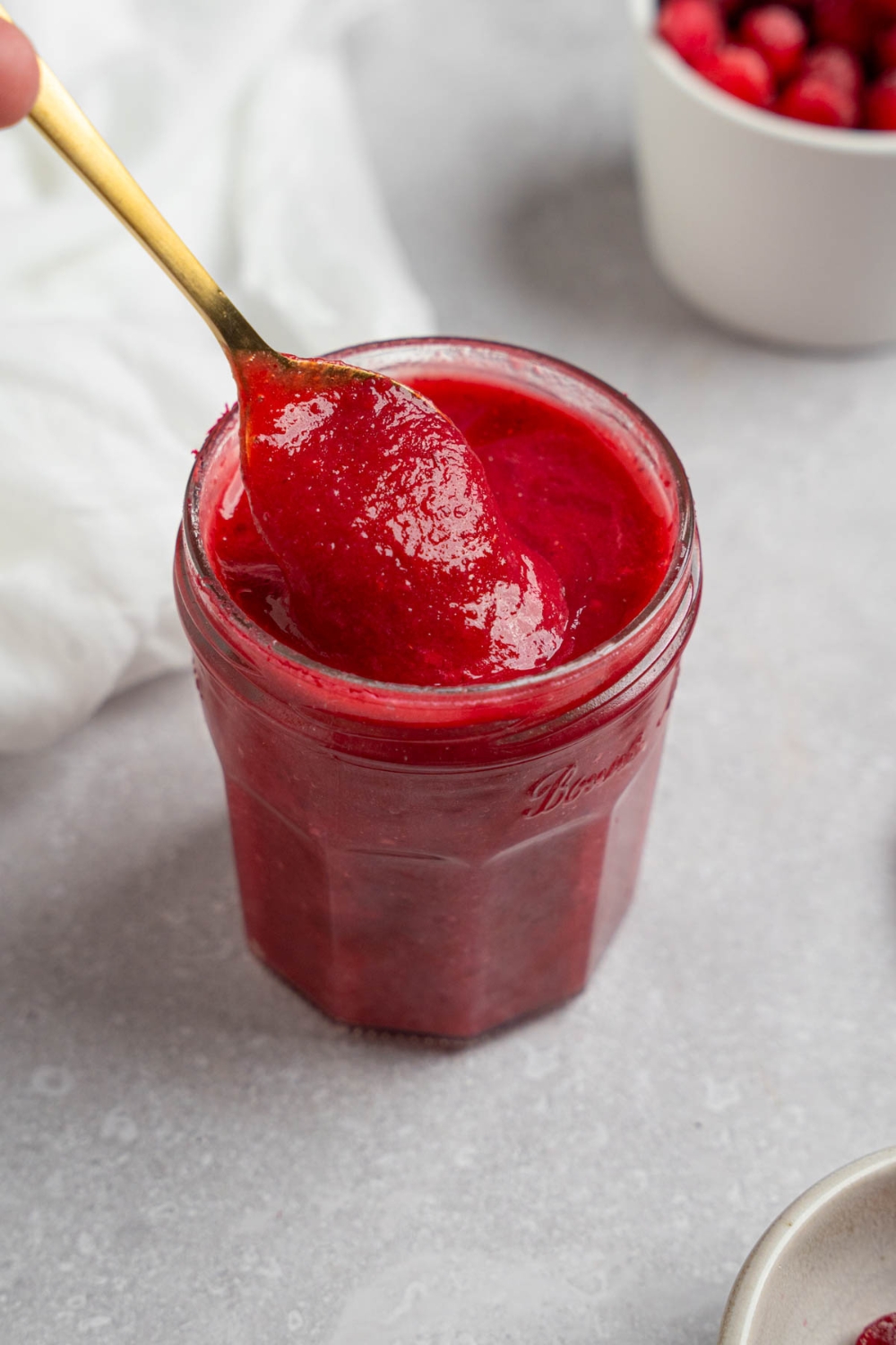A glass jar with chokecherry pudding. A spoon is stirring the pudding. The jar is on a white counter with a white cloth napkin and bowl of chokecherries.