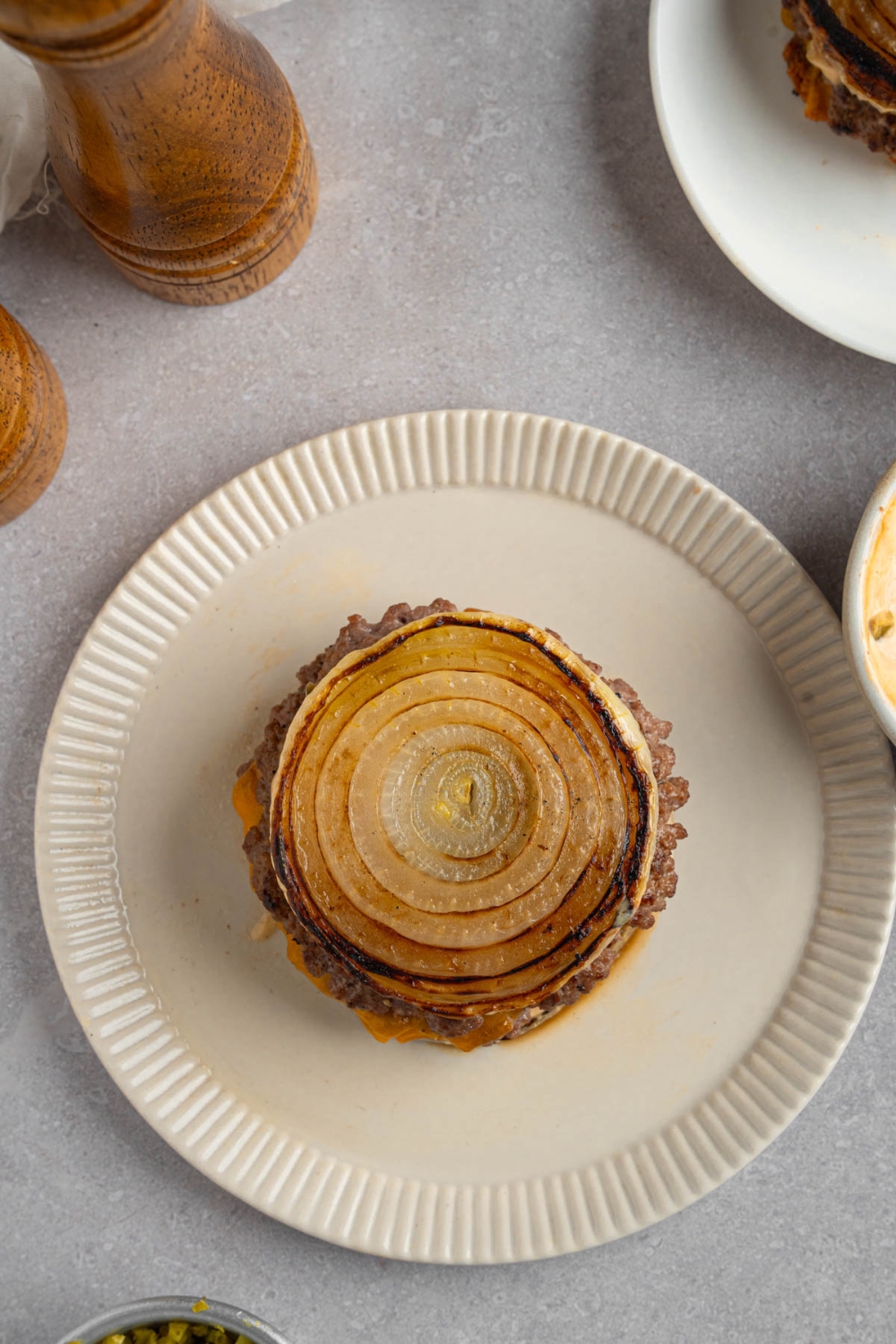 A Flying Dutchman burger in between grilled onions on a white plate. The plate is on a white counter with a pepper mill.