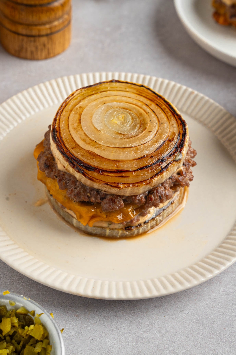 A Flying Dutchman burger in between grilled onions on a white plate. The plate is on a white counter with a pepper mill and small bowl of garnishes.