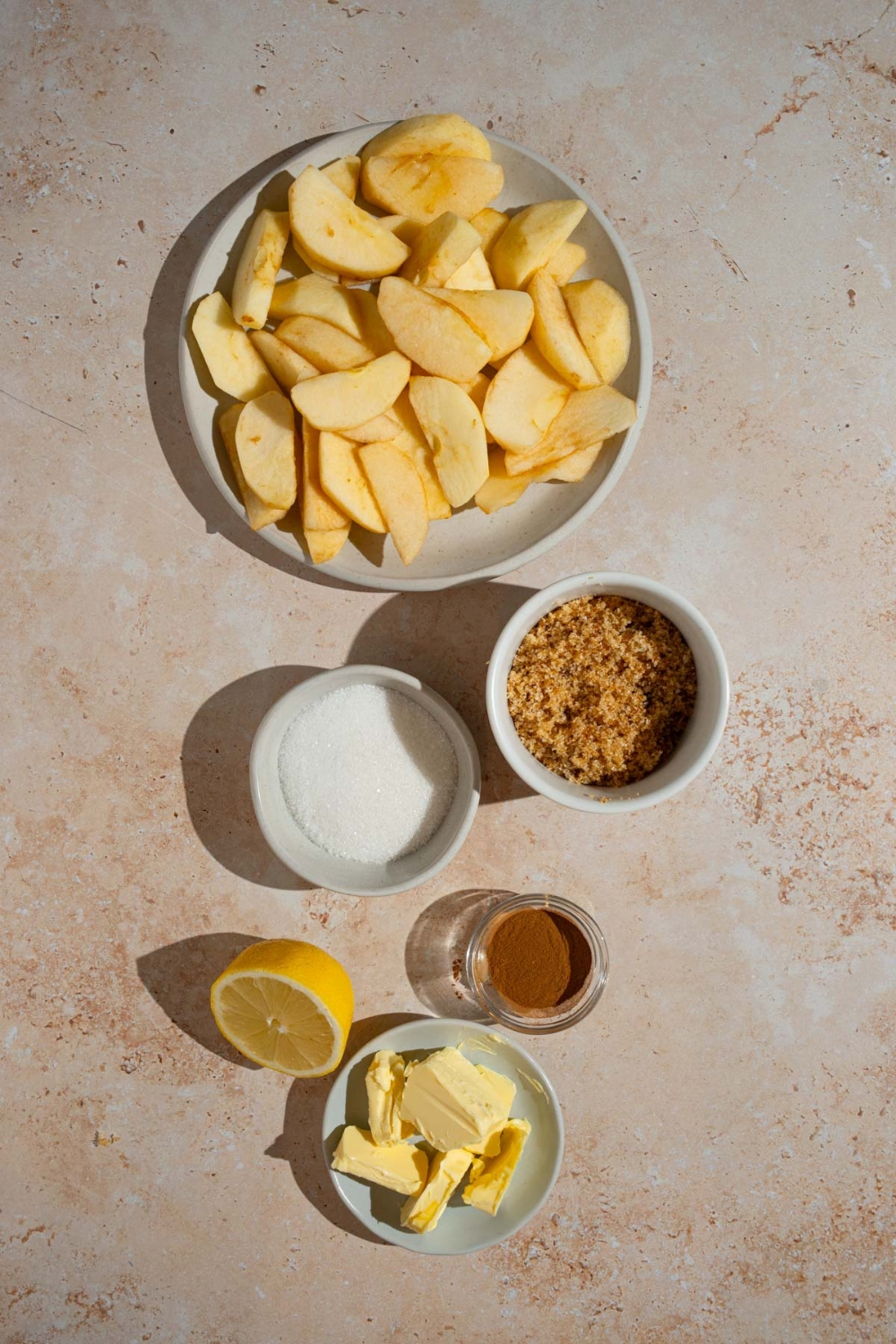 An overhead shot of several bowls in various sizes containing ingredients to make copycat Cracker Barrel fried apples including sliced apples, butter, white sugar, brown sugar, cinnamon, and lemon.