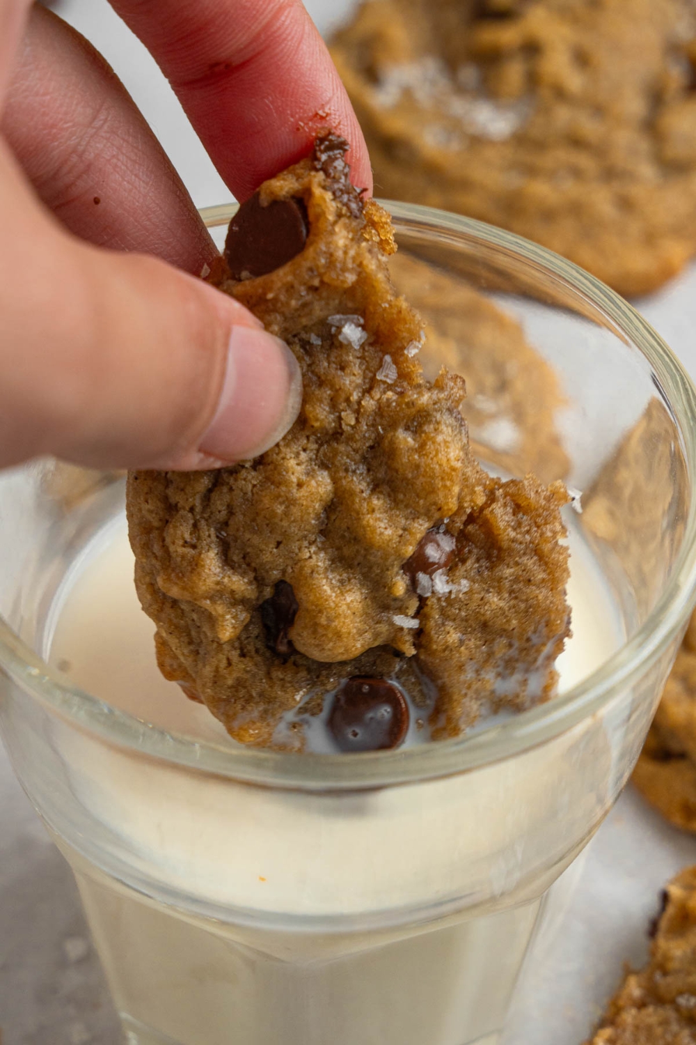 A hand dipping a bite of a banana bread cookie into a glass of milk.