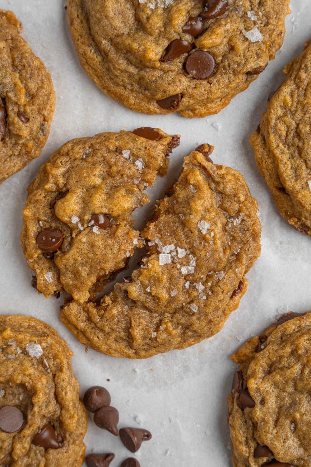 Banana bread cookies sprinkled with flaky sea salt on a baking sheet lined with parchment paper. The baking sheet is garnished with chocolate chips.
